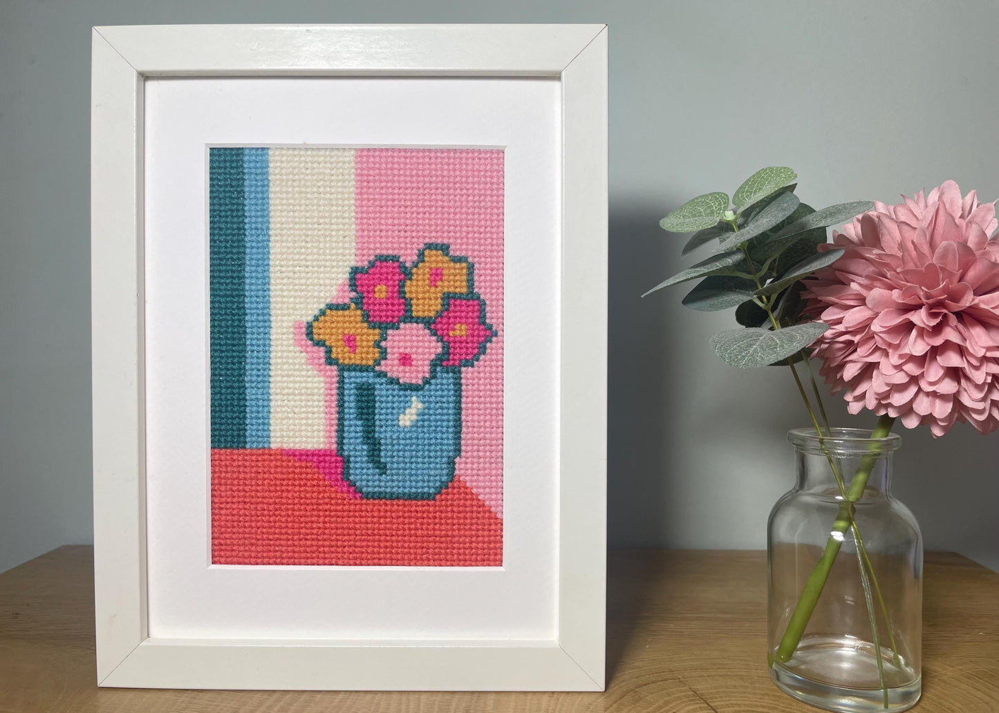 The image shows a 5 x 7 cross stitch of a pot of pink and mustard flowers sitting in a corner on a coral table against pink and white walls in a corner.